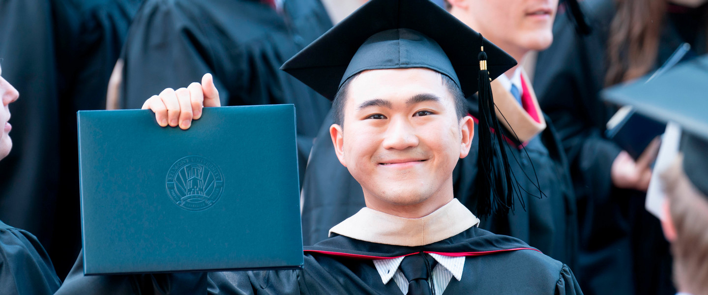 A graduate student in a cap gown and hood holding up his diploma after graduation