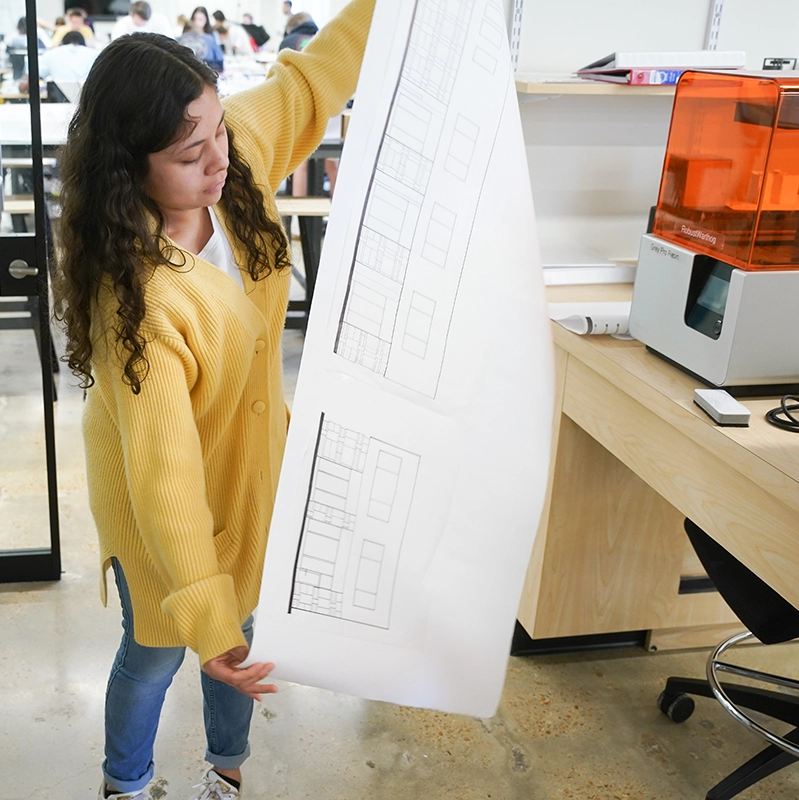 Girl standing in Digital Fabrication Lab holding a printed blueprint of a model with fabrication machine in background