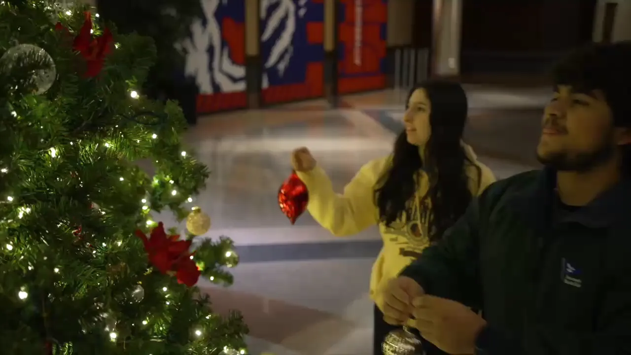 Two students are decorating a brightly lit Christmas tree with ornaments in a festive indoor setting.