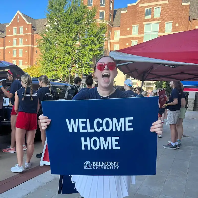 A volunteer wearing red-tinted sunglasses and a navy T-shirt stands in front of a brick campus building and holds a large blue sign that reads “WELCOME HOME” above the Belmont University logo. Behind her, other volunteers in similar attire work under pop-up tents, creating a festive move-in or welcome event atmosphere.