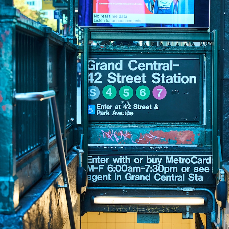 A New York City subway entrance sign for Grand Central-42 Street Station, featuring colored circles for lines S, 4, 5, 6, and 7. The sign includes accessibility information and operating hours