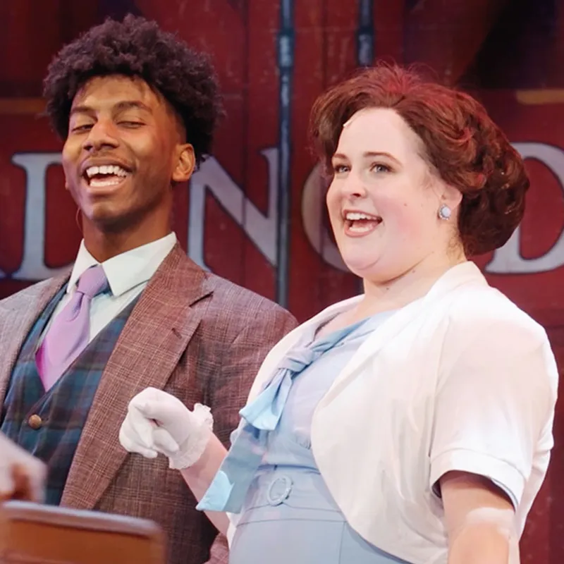 Two theater performers, one man and one woman, smiling during a performance.