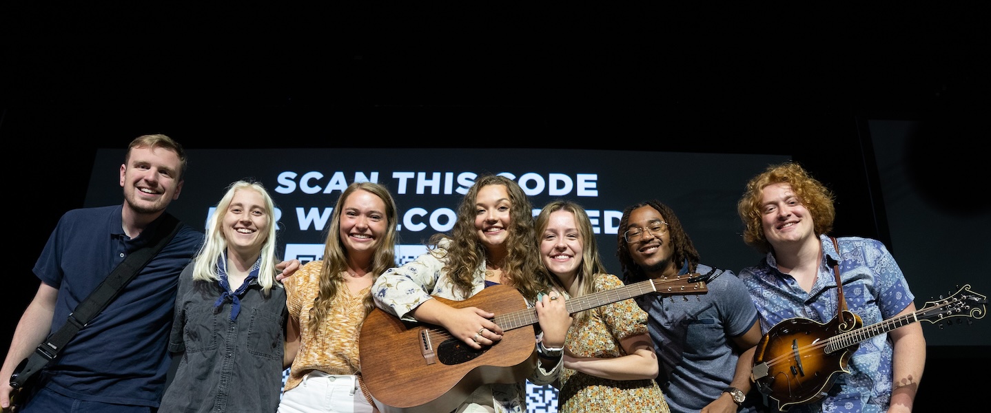 A group of seven smiling young adults stand closely together on stage, two holding instruments—a guitar and a mandolin. A black screen behind them displays white text reading "SCAN THIS CODE" with a large QR code partially visible.