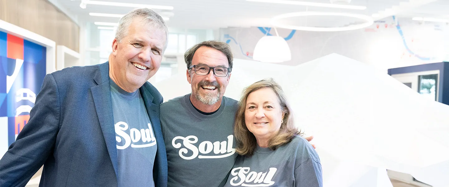 Greg Jones, Sam Simpkins, and Susan Jones standing together in the Belmont University Admissions Welcome Center, all smiling and wearing matching gray "Soul" t-shirts.