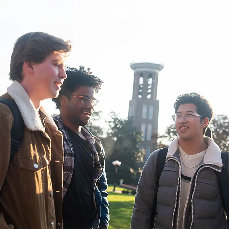 Three students stand together outdoors on Belmont University's campus, smiling and engaged in conversation. The iconic Bell Tower is visible in the background, framed by bright sunlight.