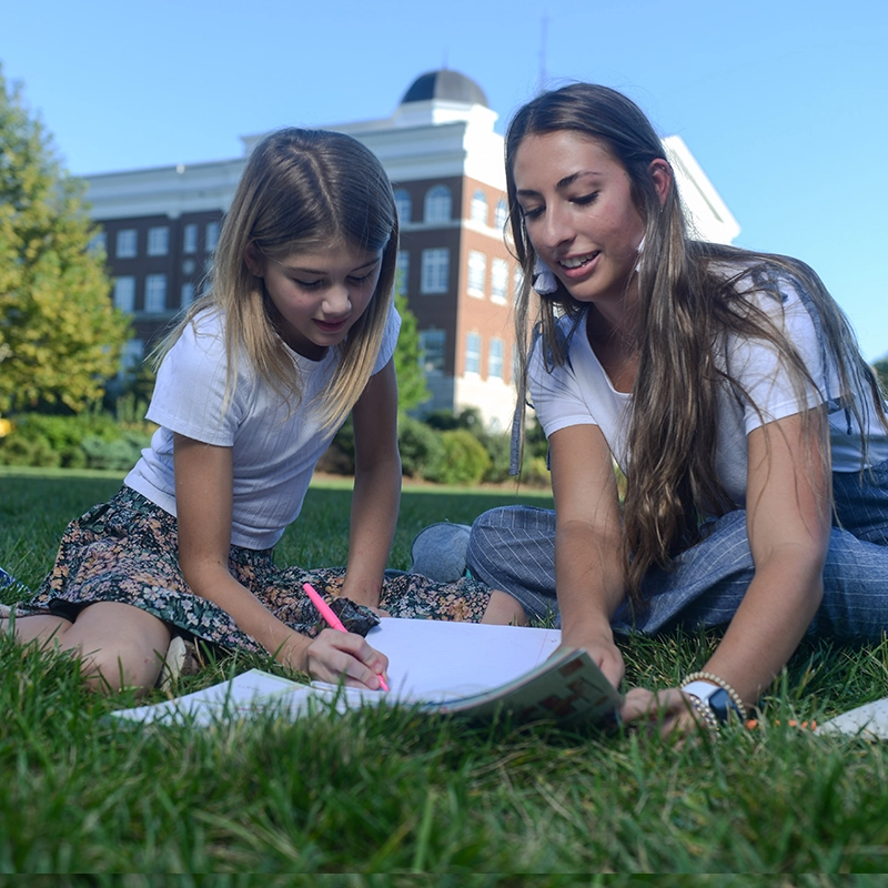 A college student and a young girl sit on the grass outdoors, sharing a notebook. The student is helping the girl with a drawing as they enjoy a sunny day on campus, with a building visible in the background.