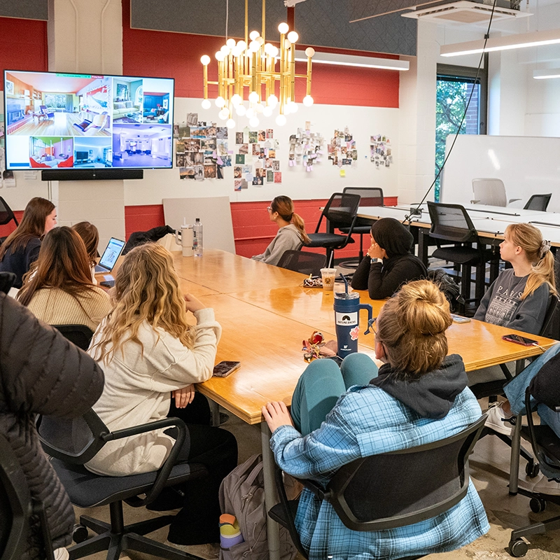 Students sit around a large wooden table in a modern classroom, attentively watching a presentation on a wall-mounted screen displaying interior design images. The room features bright lighting, a gold chandelier, red and white walls, and a collage of inspiration photos on the back wall.