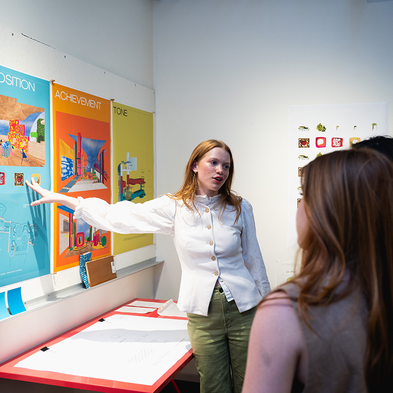 A student in a white blouse and green pants presents her colorful design project to two peers, pointing to a display board labeled “COMPOSITION,” “ACHIEVEMENT,” and “TONE” in a well-lit interior design studio. Drafts and materials are laid out on a red table beneath the board.