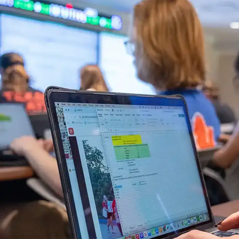 Students are engaged in a classroom setting, focusing on their laptops during a lecture. One student is seen typing, with a spreadsheet displayed on the screen, while large monitors in the background show relevant data. The atmosphere is collaborative and academic, highlighting the learning experience in the MBA and MACC programs.