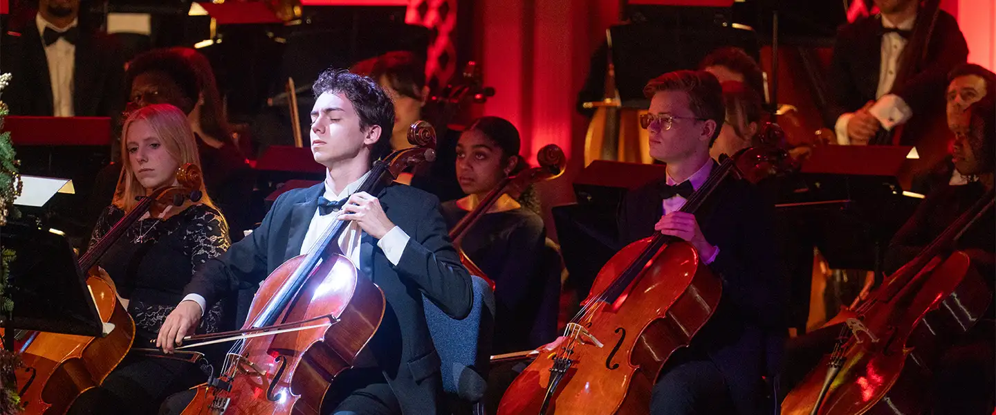 A group of musicians in formal attire performing in an orchestra, with a focus on two cellists. One cellist is deeply engrossed in playing, while a female musician is seated nearby, looking on attentively.