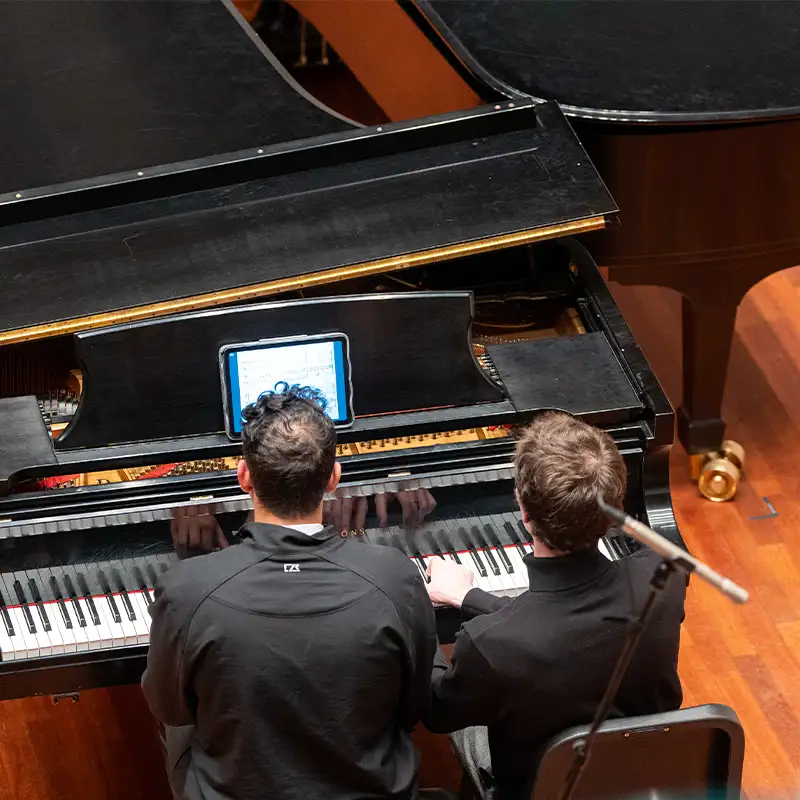 Two pianists are seated at a grand piano, playing together. From a top-down perspective, their hands are visible on the keys, and an iPad displaying sheet music is positioned on the piano. The setting features a polished wooden floor and another grand piano in the background, creating an intimate performance atmosphere.