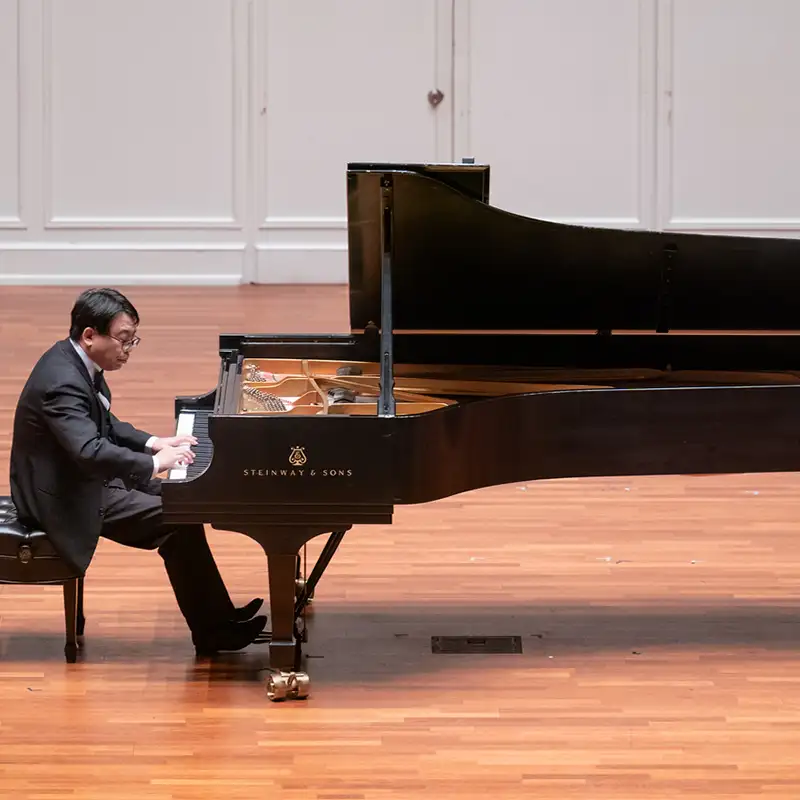 A pianist in a black suit is performing at a grand piano on a polished wooden stage. The piano is a Steinway & Sons model, and the backdrop features a simple, elegant design. The musician is focused on playing, with a thoughtful expression.