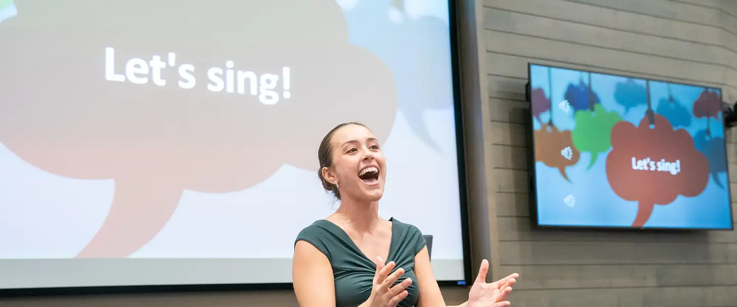 A woman with a ponytail is singing in front of a  classroom, gesturing with her hands. Behind her is a screen displaying the text "Let's sing!" in a speech bubble graphic.
