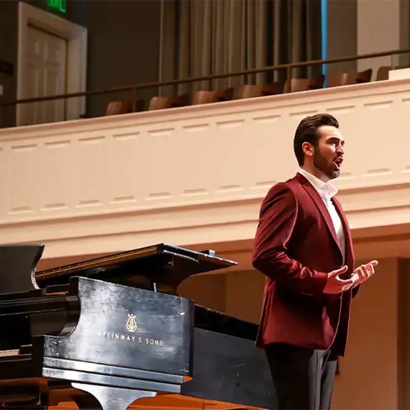 A male vocalist in a burgundy suit sings passionately on stage, with a grand piano visible in the foreground. The performance takes place in a well-lit concert hall, showcasing elegant architecture and seating. The singer is gesturing expressively, engaging the audience with his performance.