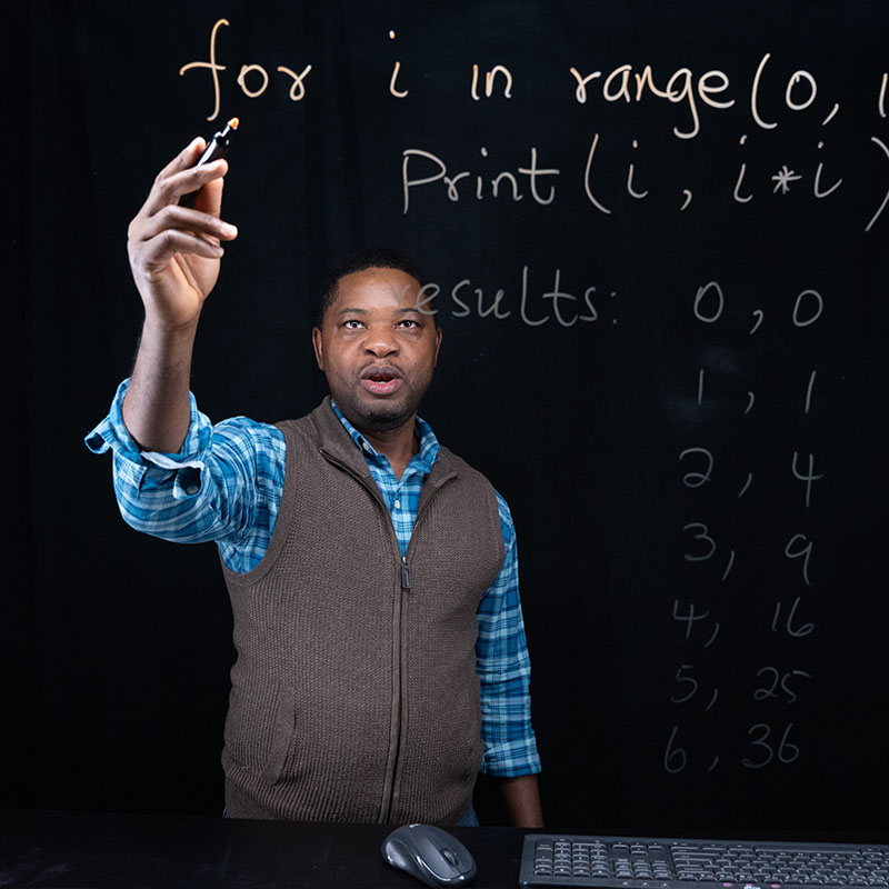 An instructor demonstrates a programming lesson, writing a Python loop and output on a glass board.