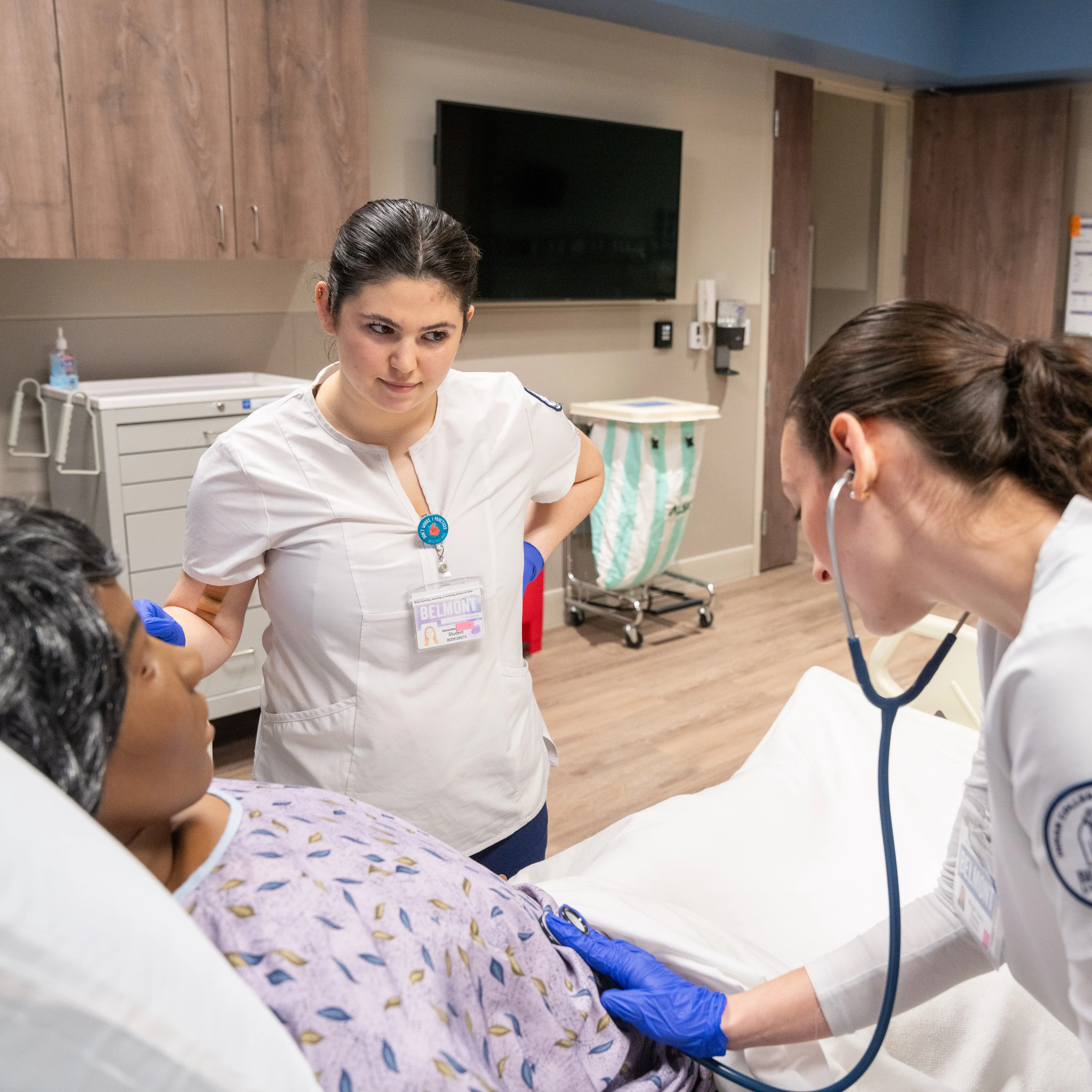 Two Belmont nursing students in scrubs work with a patient manikin in a simulation lab, one listening with a stethoscope while the other observes.