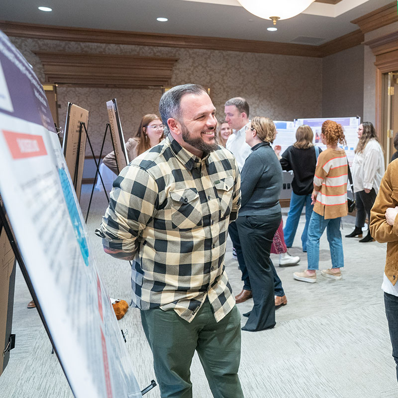 People interacting and discussing a poster at the Belmont Social Work Symposium, with a man in a checkered shirt smiling in the foreground.