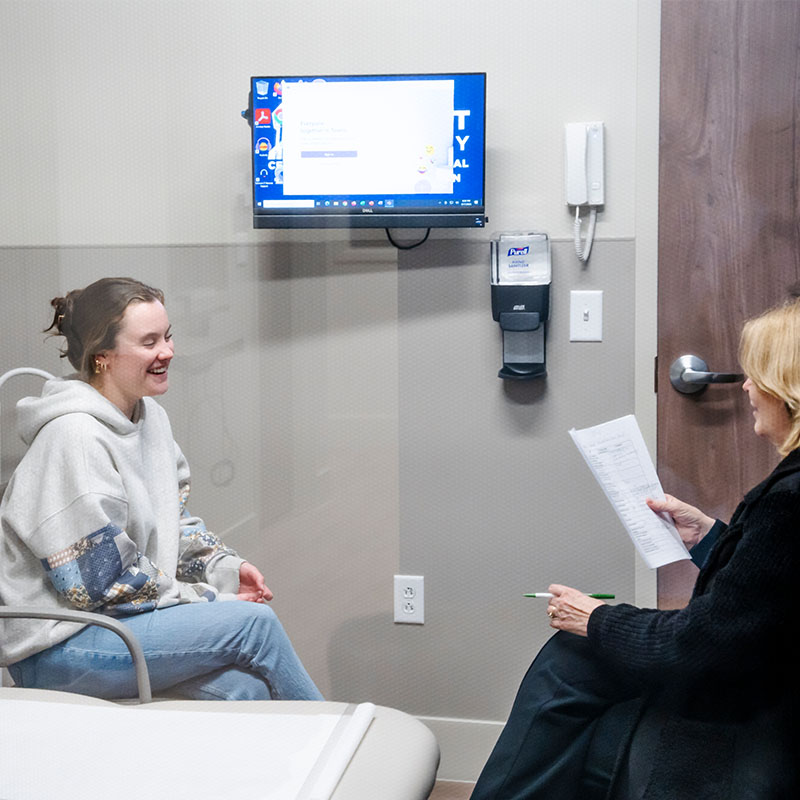 A student laughs during a simulation session with an instructor holding a clipboard in a medical examination room equipped with a wall-mounted computer screen