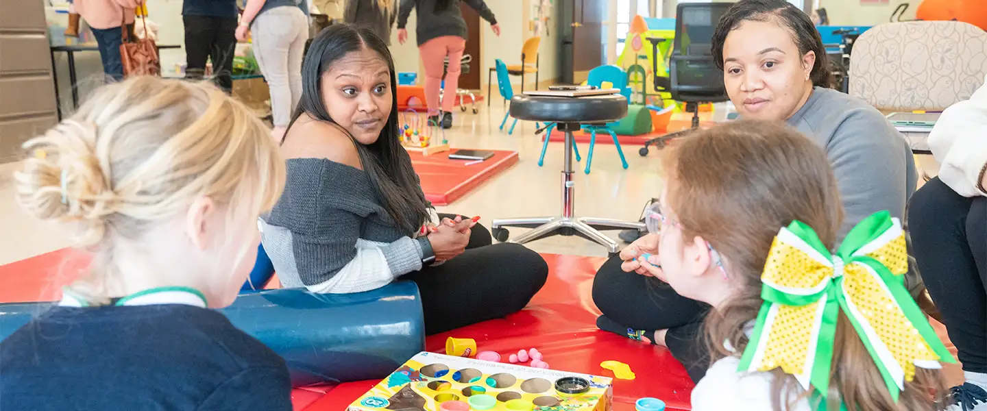 An indoor photo of two Occupational Therapists seated and engaging with pediatric patient