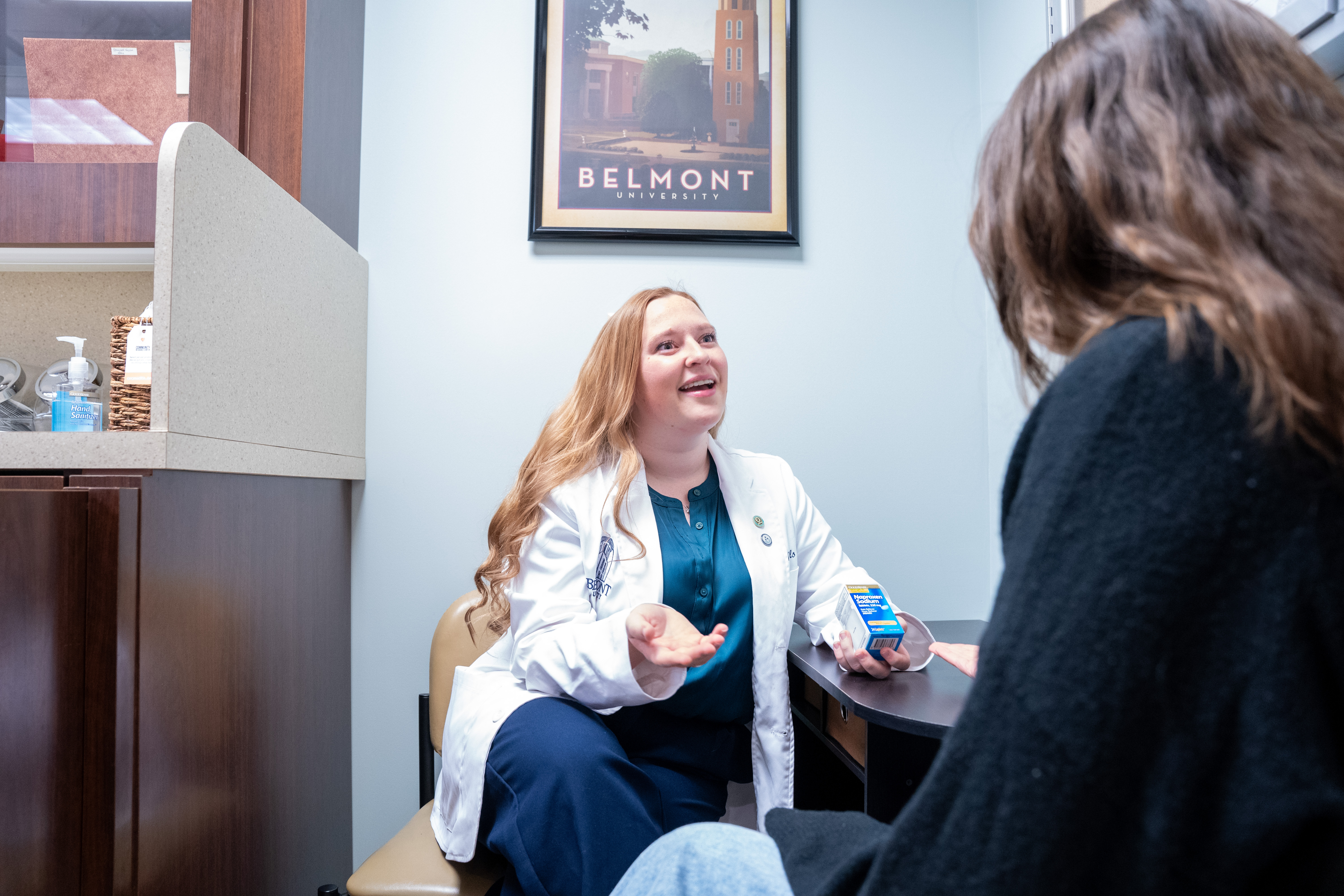 A female healthcare professional in a white lab coat sits at a desk, smiling and gesturing as she speaks to a patient. The patient, partially out of frame, has long brown hair and is wearing a black sweater. In the background, there is a poster of Belmont University on the wall, and a small basket containing supplies is visible on the desk.