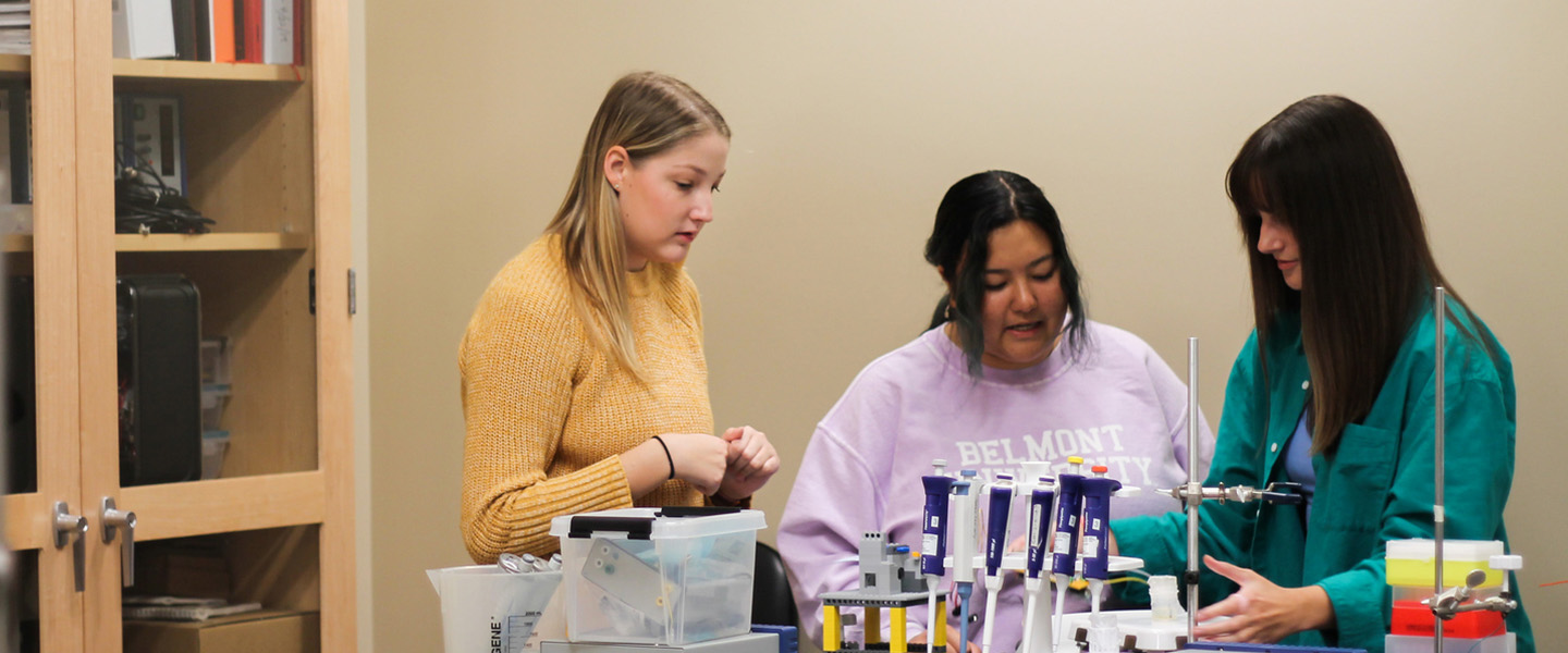 Three female students stand behind a lab table and look down at a series of metal instruments.