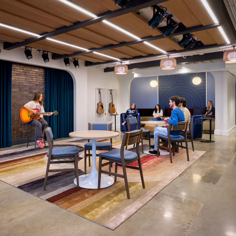 A person plays guitar on a small stage with a blue curtain backdrop. An audience of five listens attentively in a modern cafe setting with wooden tables.