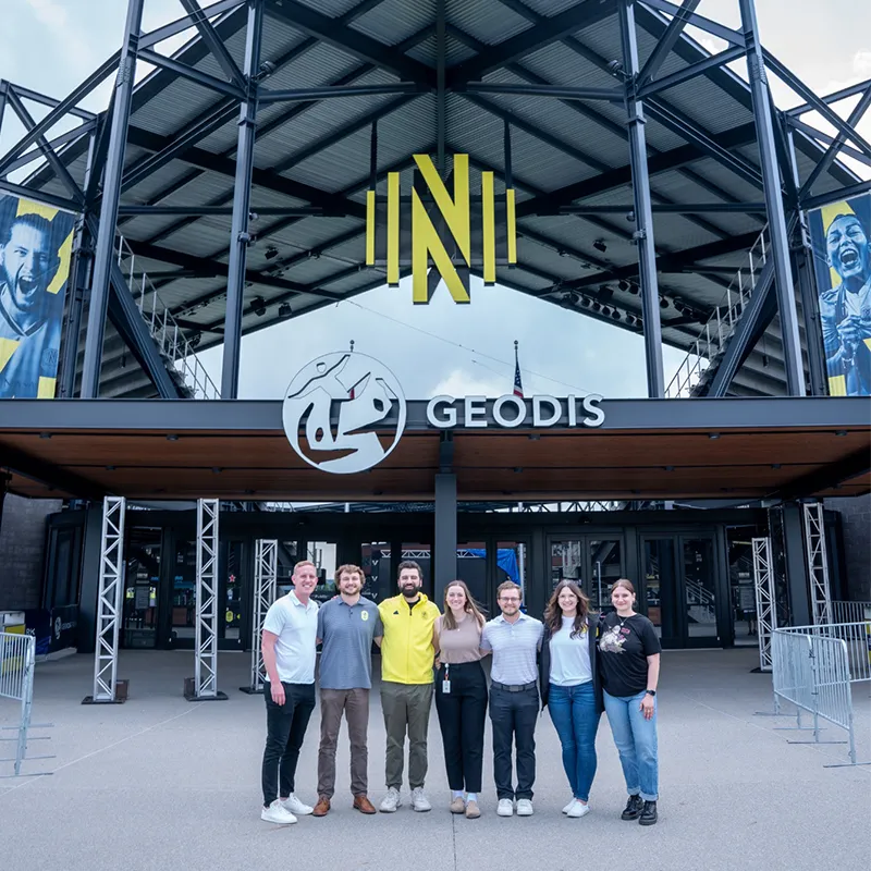 A group of Belmont University graduates stands in front of the Geodis Park entrance, home of Nashville SC, with the club's iconic yellow logo prominently displayed above them.