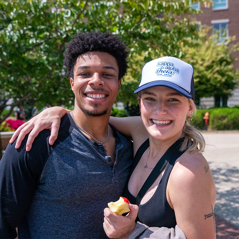 Two smiling students standing outside, one with a Kappa Alpha Theta cap, the other holding an apple, enjoying a sunny day on campus.