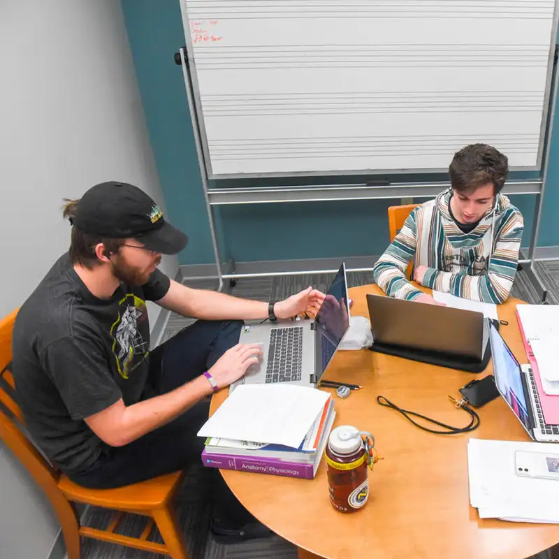 Two students sit at a table with laptops and study materials, working together in a study room with a whiteboard behind them.