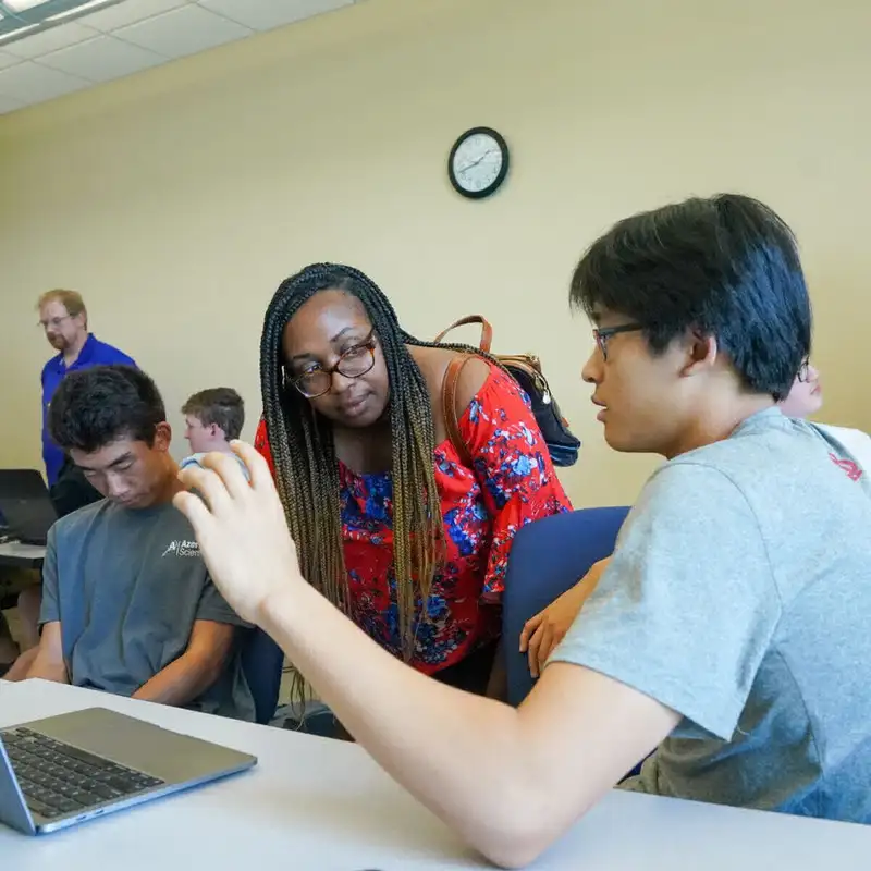 An instructor leans over to talk with students during a workshop or class, as they work on laptops in a classroom setting.
