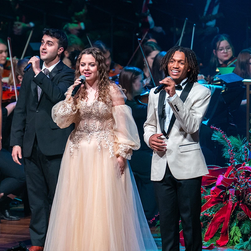 Three Belmont University students perform a vocal piece during a formal concert. The singers, dressed in elegant attire, hold microphones and sing in harmony.