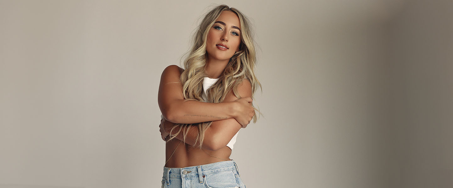 Country artist Ashley Cooke poses confidently against a neutral background. She has long, wavy blonde hair and is wearing a casual, sleeveless white crop top and light-wash denim jeans. Her arms are crossed gently over her chest, and she gazes directly at the camera with a soft yet empowered expression.