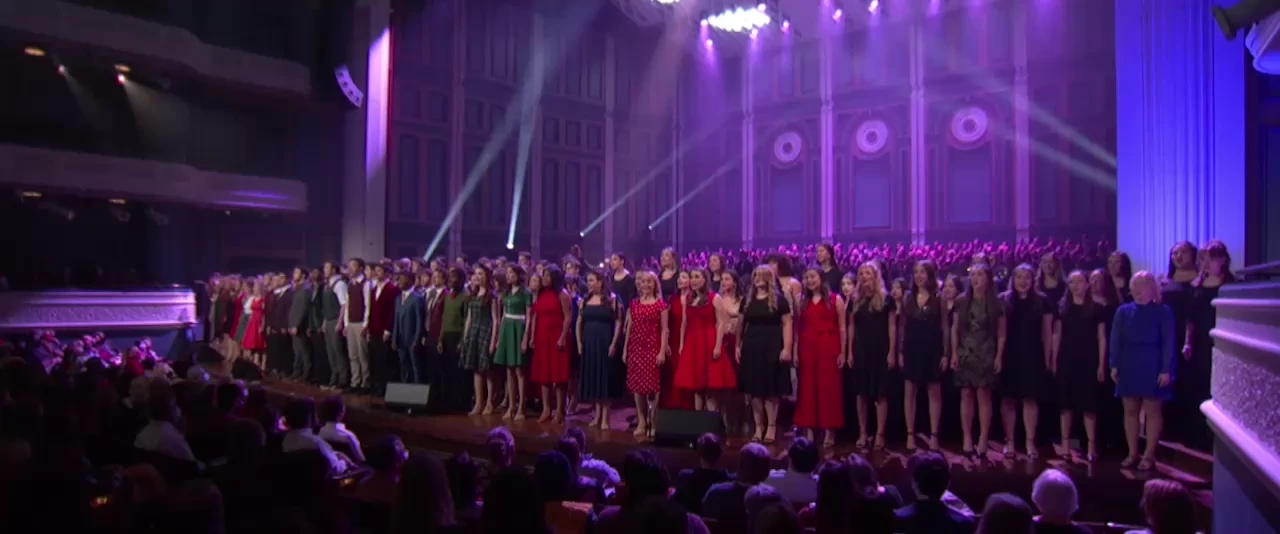 A large choir of Belmont University students performs on stage during the Christmas at Belmont concert, held in the Fisher Center for the Performing Arts. The singers are dressed in festive attire, with a mix of red, green, and black dresses and suits. They stand in a long row at the front of the stage, illuminated by vibrant stage lights.