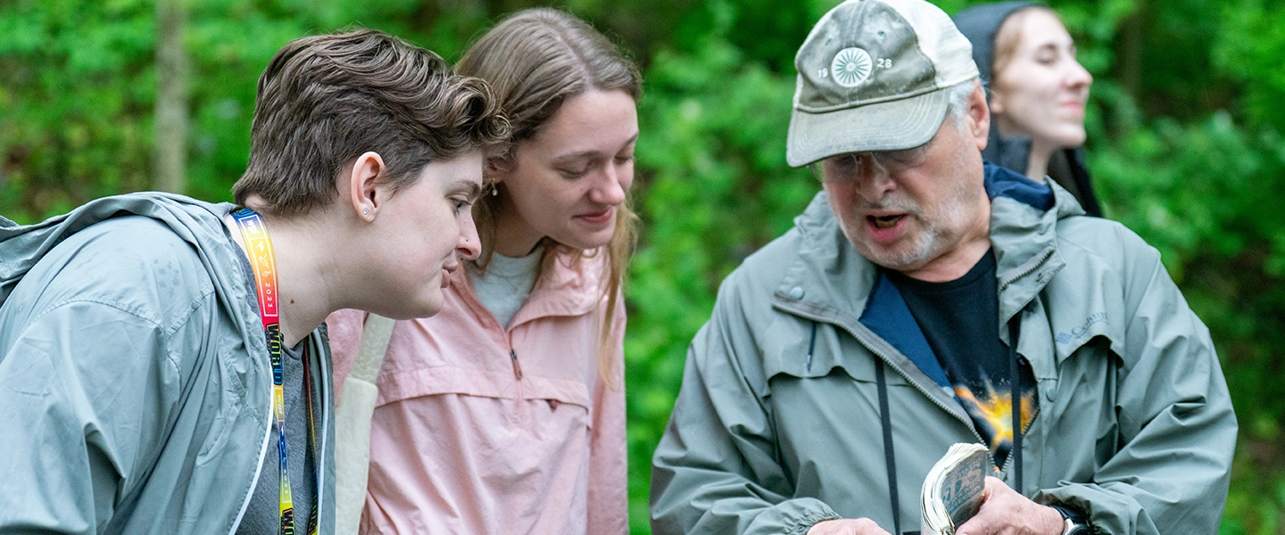 While outdoors among trees, an older man showing a book to two young people.