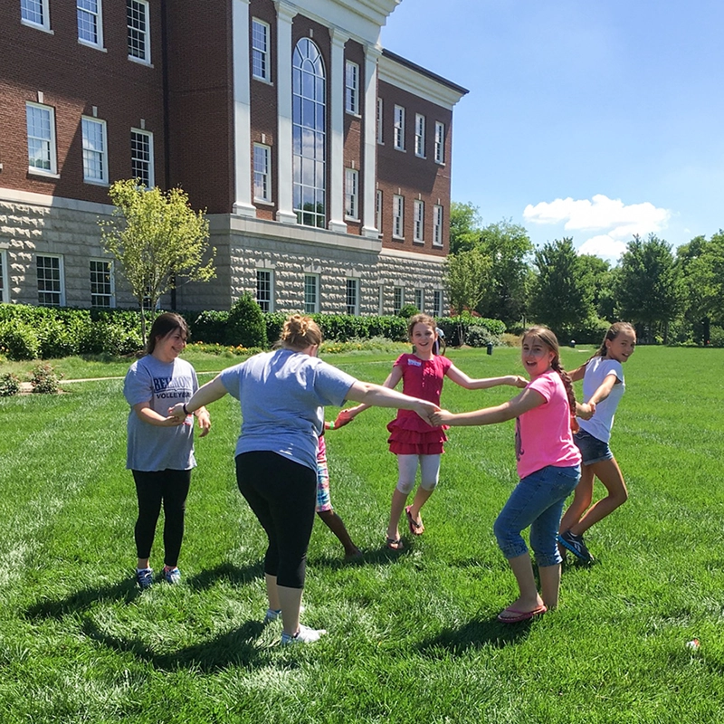 Kids and Belmont Students hold hands in a circle smiling on the lawn