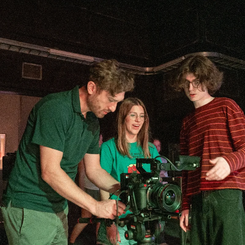 Three students gather around a professional video camera on a tripod in a dim studio space, adjusting settings and reviewing the equipment together.