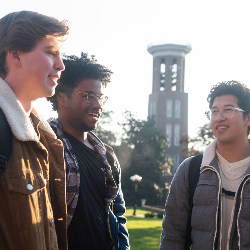 Three Belmont University students talking and smiling outdoors on campus, with the iconic Bell Tower visible in the background during sunny weather.