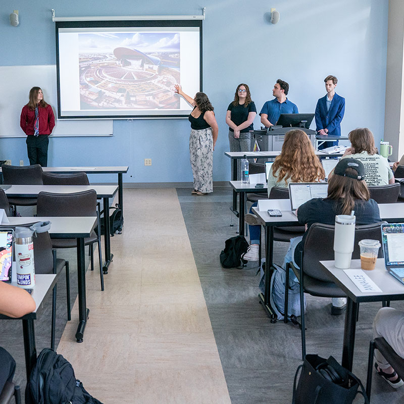 Students in a Belmont University classroom presenting a group project to their peers, with one student pointing to a projected image of a stadium on a screen