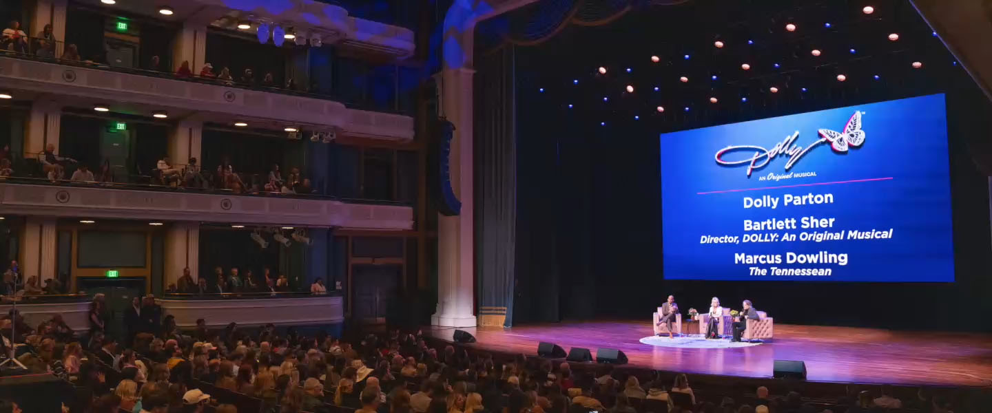 A wide-angle view of the Fisher Center at Belmont University during a special event for Dolly: An Original Musical. The stage features a large screen displaying the musical's logo and the names of panelists: Dolly Parton, director Bartlett Sher, and The Tennessean's Marcus Dowling. The audience fills the theater, and the panelists are seated in a discussion onstage under warm stage lighting.