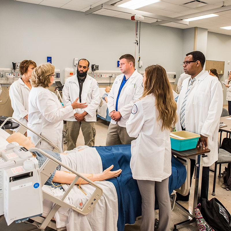 Professor speaking to students while they stand around a hospital bed with a medical dummy in simulation lab
