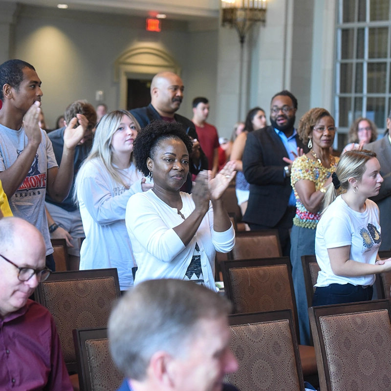 A diverse group of people standing and applauding during an event in a large, elegant room with ornate lighting and tall windows. Their expressions reflect enthusiasm and appreciation.