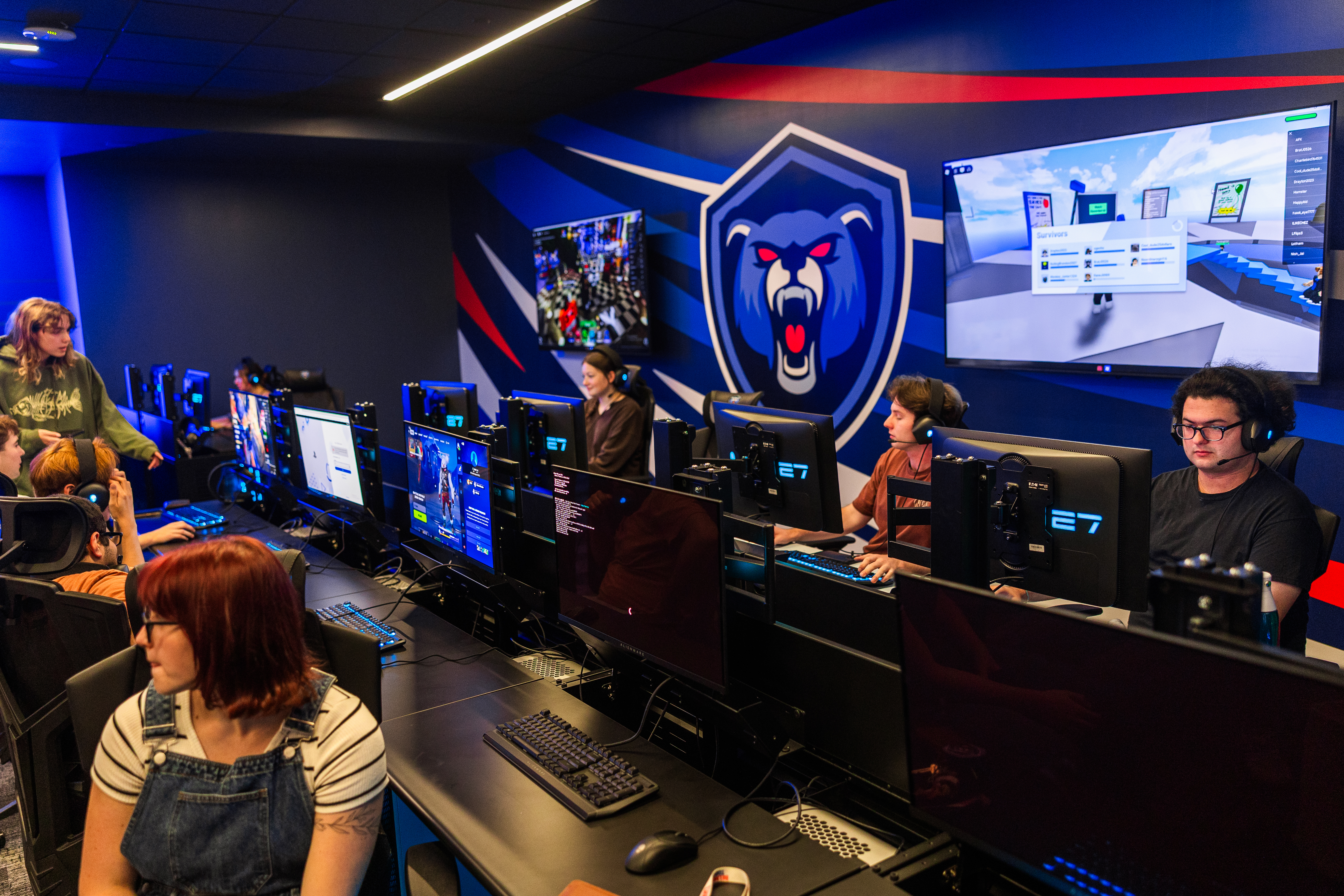 Rendering of an esports space with teams seated at computers, a leaderboard on the wall, and screens displaying match graphics.