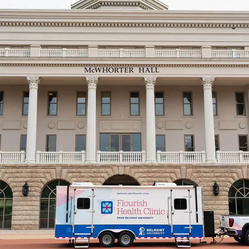 A mobile health clinic is parked in front of McWhorter Hall, a stately building featuring columns and large windows. The clinic is branded as "Flourish Health Clinic" from Belmont University, showcasing a colorful design. The image captures the clinic alongside the building, emphasizing its role in providing healthcare services.