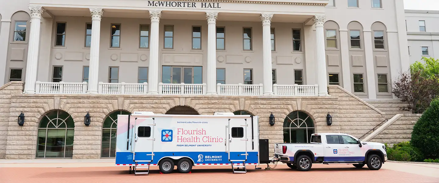 A mobile health clinic is parked in front of McWhorter Hall, a large, elegant building with columns and large windows. The clinic is branded as "Flourish Health Clinic" from Belmont University, featuring a colorful design. A pickup truck is attached to the clinic, emphasizing its mobile nature.