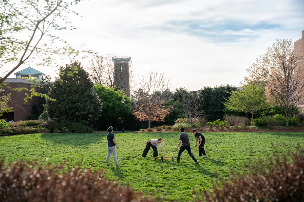 Four people play a game on a green lawn surrounded by trees and buildings under a blue sky.