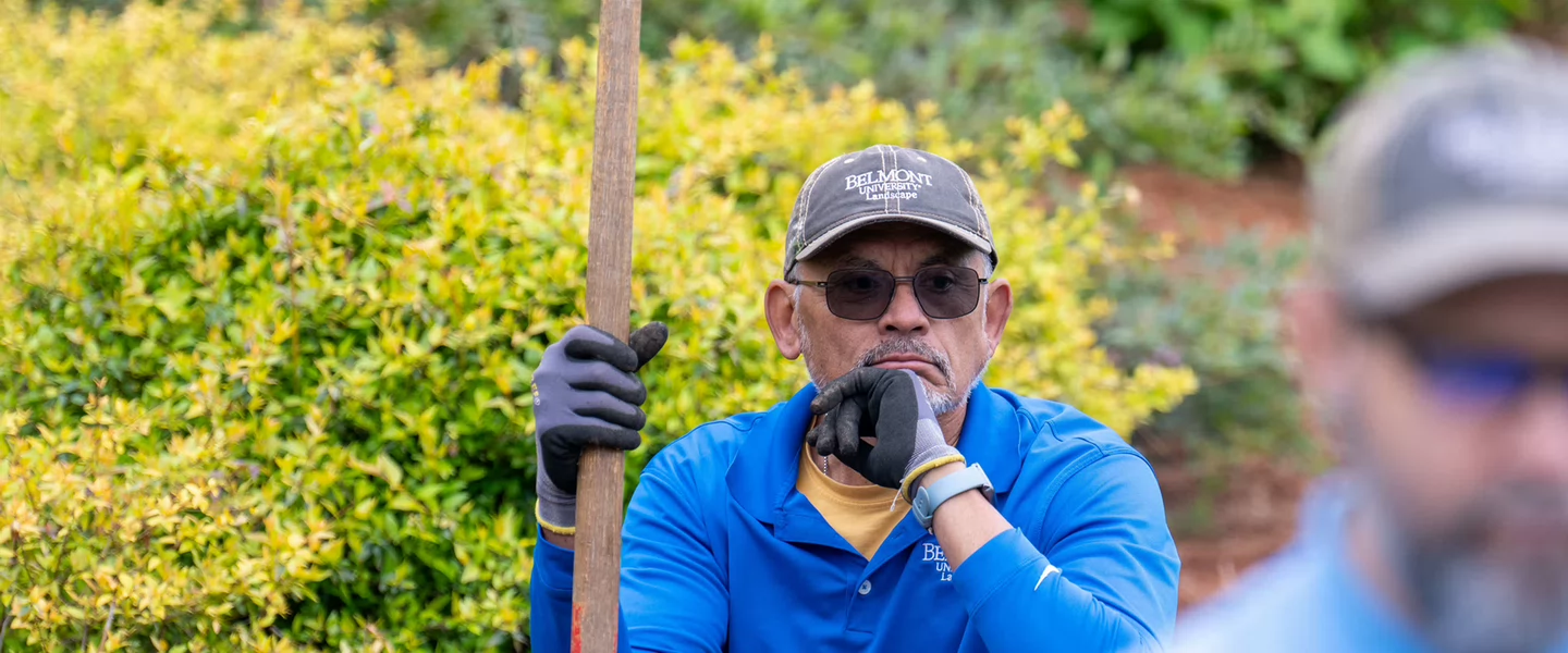 A man in a blue shirt and gloves holds a shovel, sitting pensively in a garden with leafy plants in the background. Another man is blurred in the foreground.