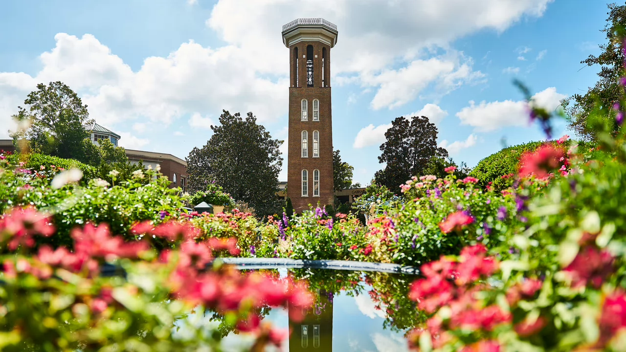 A tall, brick tower stands against a blue sky, surrounded by a vibrant garden of pink and purple flowers. A pond reflects the scene.