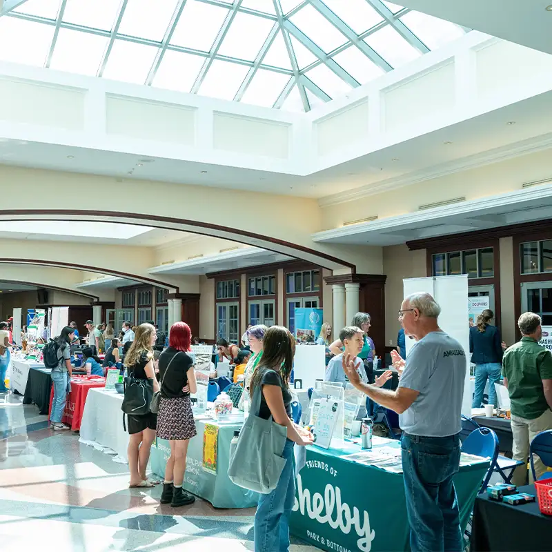 A bustling indoor event hall filled with people at tables displaying various informational booths. Sunlight streams through a large skylight, illuminating attendees who are engaged in conversation and examining materials. In the foreground, tables feature colorful banners and brochures, while groups of people interact throughout the space.