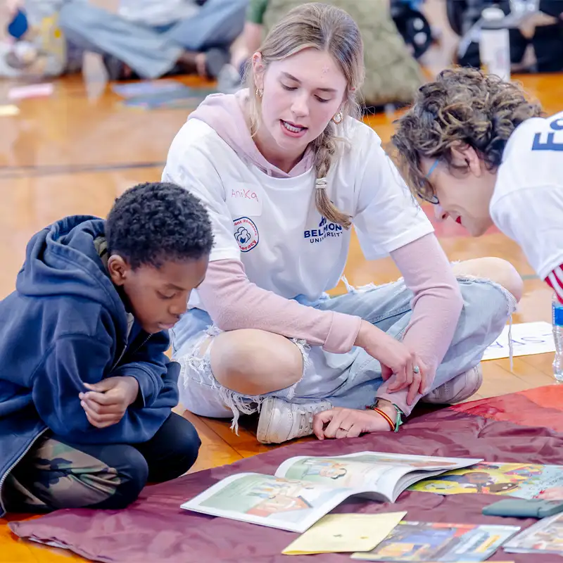 A young child sits on the gym floor, focused on a colorful storybook in front of him, while a college student in a white t-shirt engages him, pointing at the pages. Another individual, also dressed in a white t-shirt, leans in to listen. The setting is a lively indoor space, with other participants scattered in the background, fostering a warm and educational atmosphere.