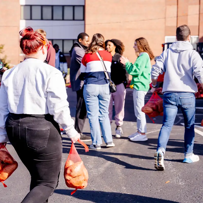 A person with bright red hair walks away from a gathering, carrying two bags of sweet potatoes in red mesh bags. In the background, a diverse group of individuals can be seen engaging in conversation, some also holding bags of produce. The setting appears to be an outdoor community event, with a building and stacks of bags visible in the background.